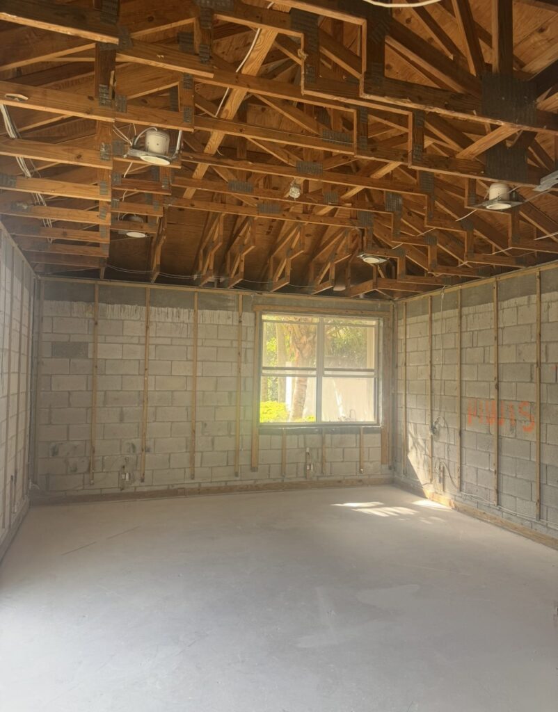 Room taken back to bare block walls and open roof trusses, with drywall, insulation, and ceiling removed in a full interior strip out.