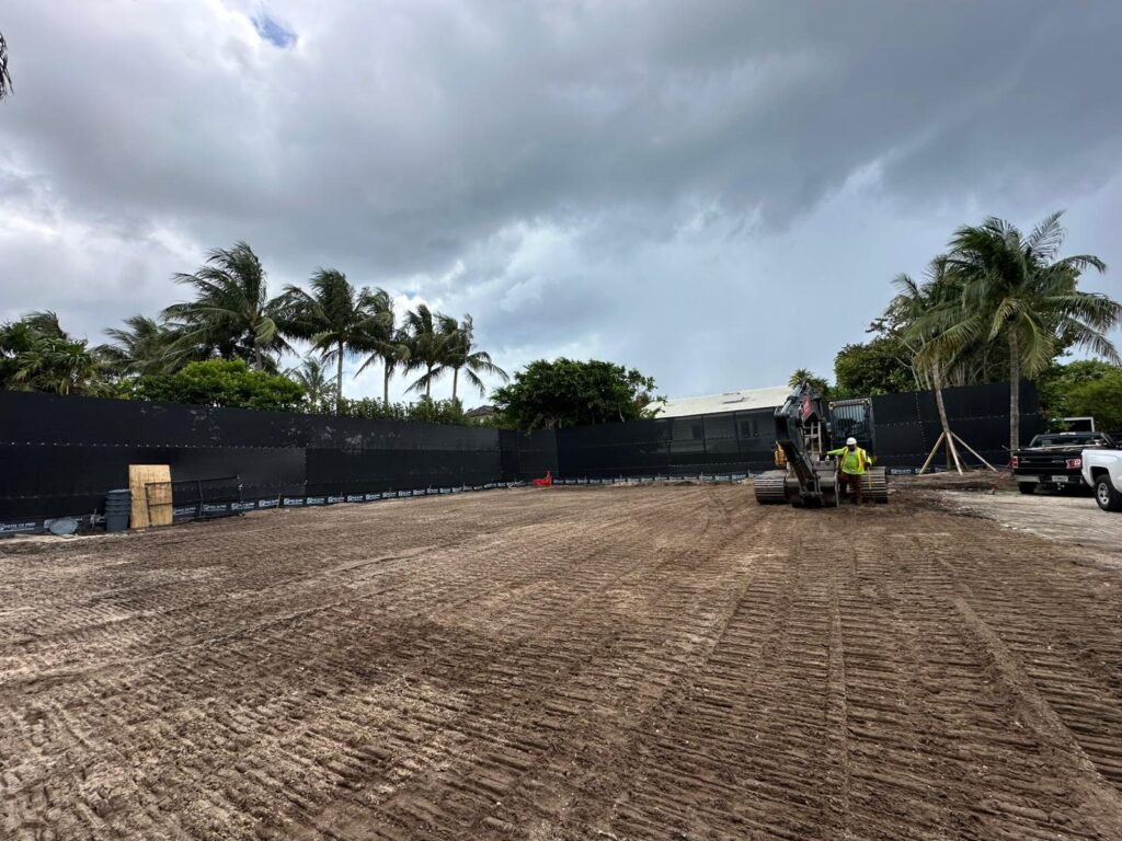 Excavator finishing grade on a cleared residential lot with privacy fencing installed around the full perimeter.